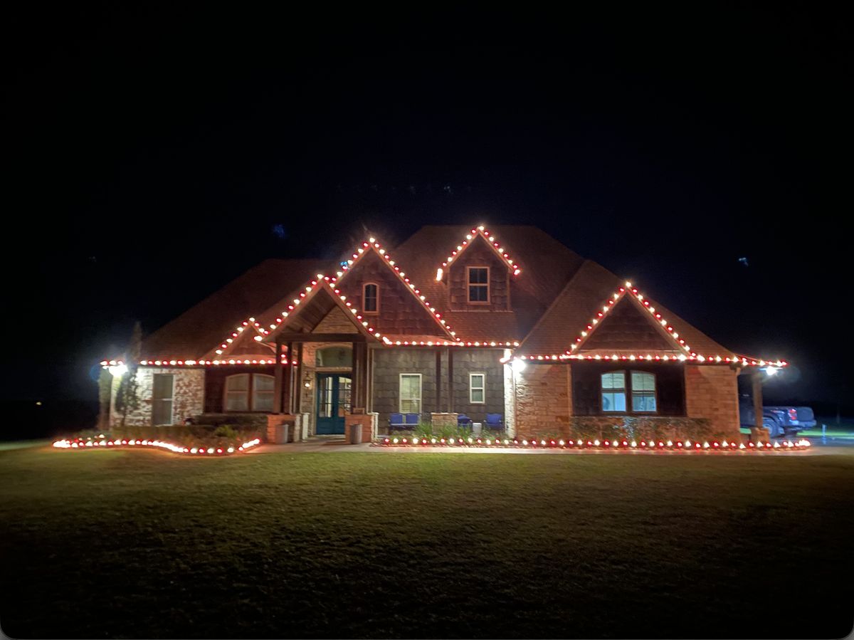Crisp warm-white bulbs following the steep gable rooflines and eaves of a large brick home against the night sky